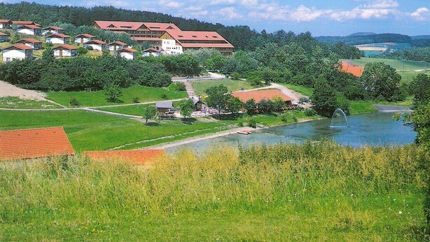 A rural village with houses, a river, and a large building with a red roof. - Bungalow 23 for up to 4 people at the RHÖN RESIDENCE vacation complex. (Dipperz)