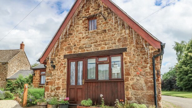 Uma casa de pedra com porta de madeira, uma pequena janela acima da porta e uma placa com a inscrição 'Smithy Cottage'.