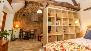 Quarto rústico com vigas de madeira aparente, parede de pedra, estante de madeira, área de jantar com mesa e cadeiras, e cama com colcha florida.