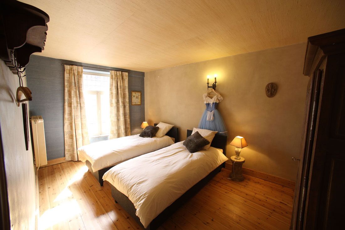 A room with two beds, a window with curtains, a wall-mounted lamp, and a small table with a lamp.