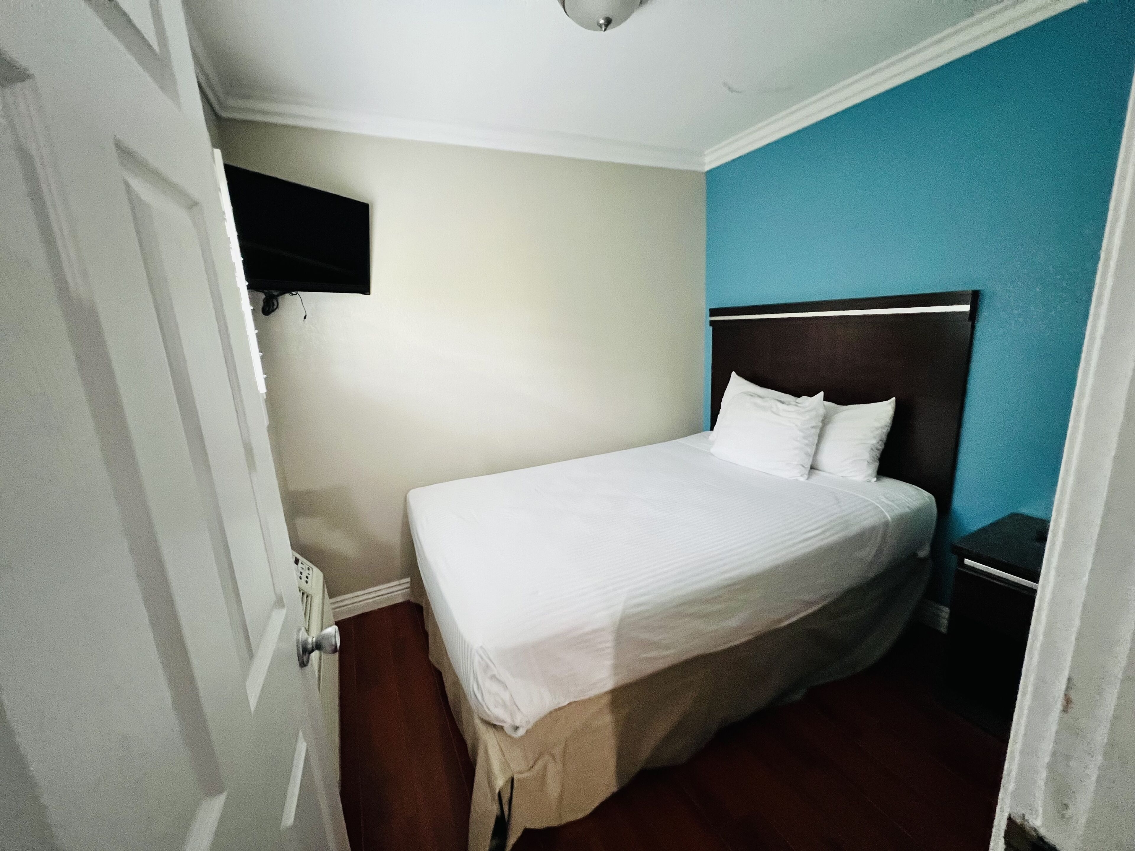 A bedroom with a single bed, a nightstand, a wall-mounted TV, and a door.
