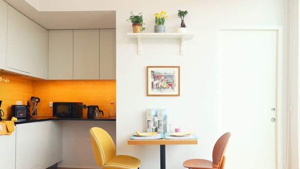 A compact kitchen with white cabinets, a microwave, and a coffee machine. There are two yellow chairs and a small table with a picture frame and a vase with flowers. - Villa Invicta II (Porto)