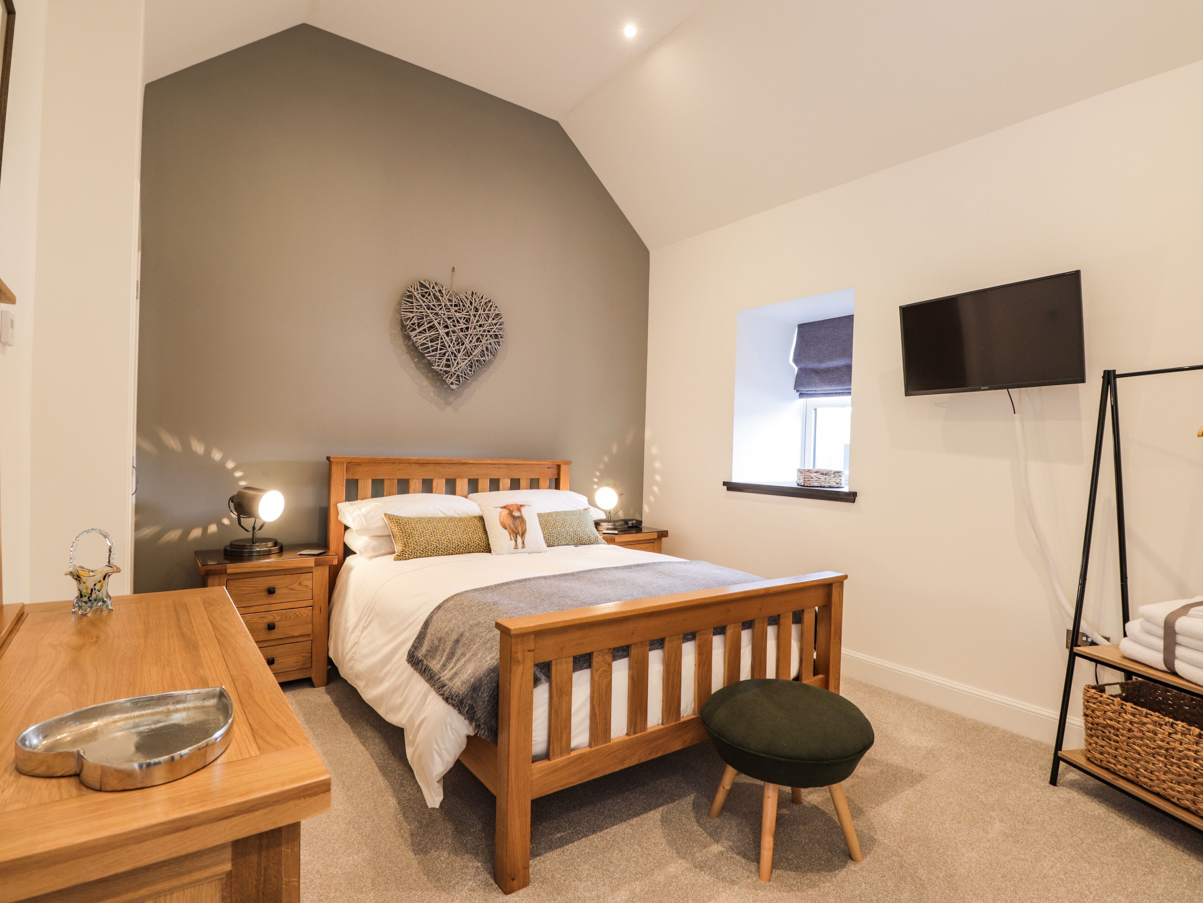 A bedroom with a sloped ceiling, a bed with a wooden headboard, a bedside table with a lamp, a small stool, a TV on a stand, and a window with a view of the outside.