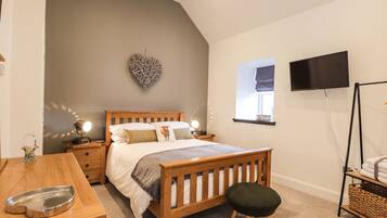 A bedroom with a sloped ceiling, a bed with a wooden headboard, a bedside table with a lamp, a small stool, a TV on a stand, and a window with a view of the outside.