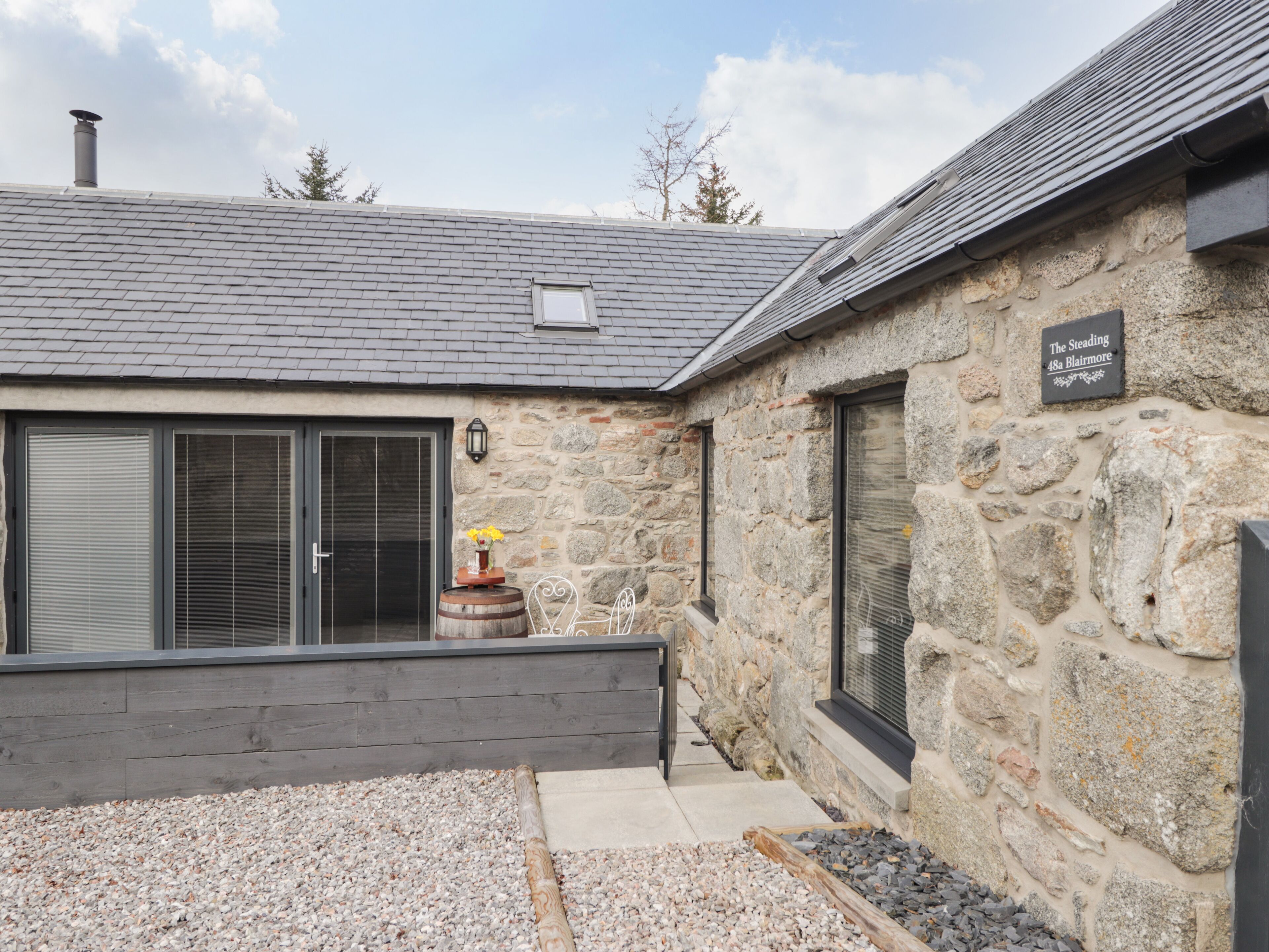 Un edificio de piedra con techo gris, un barril de madera y un letrero que dice "The Steading 48a Blairmore".