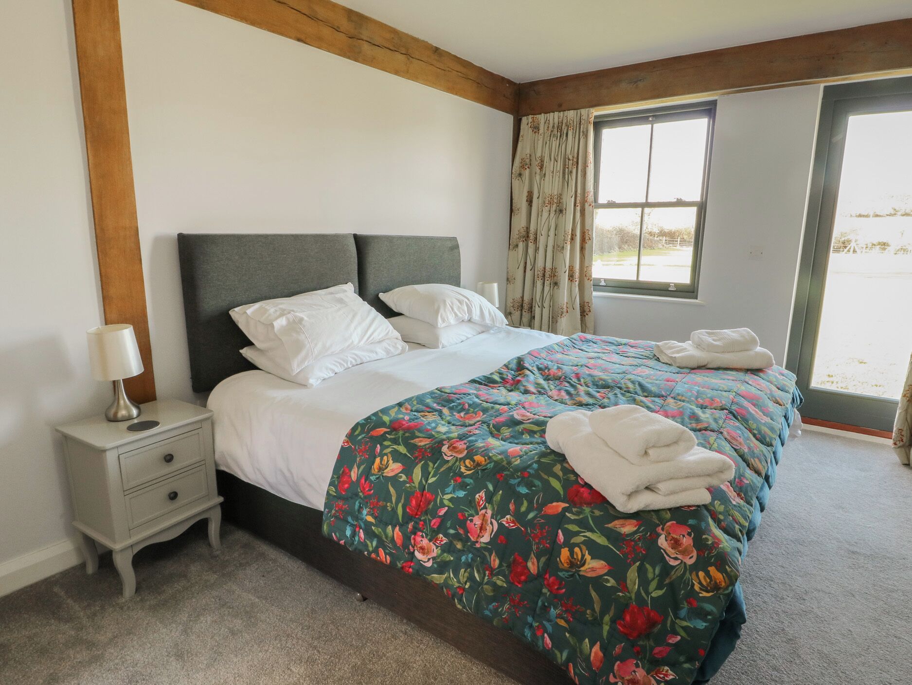 Una cama bien hecha con ropa de cama floral, una mesita de noche con lámpara y una ventana con cortinas.