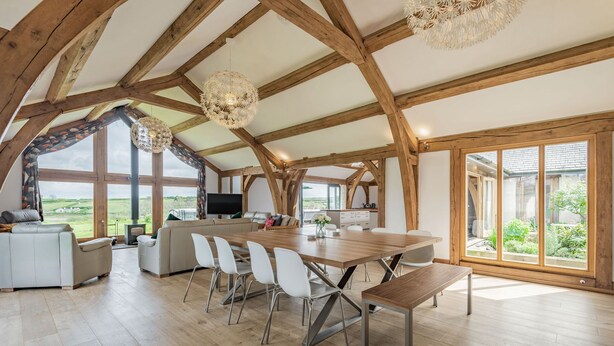 A spacious, open-plan living area with a wooden ceiling, large windows, and a central dining table with chairs.