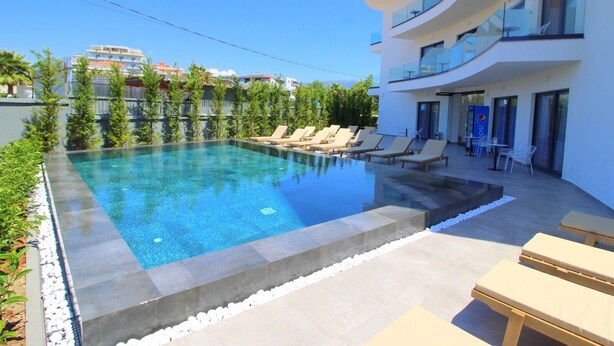 Zona de piscina moderna amb gandules i una piscina blava cristal·lina, envoltada de vegetació i un edifici blanc amb balcons.
