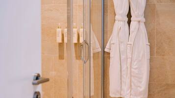 A modern bathroom with a glass shower enclosure, beige tiles, and two white robes hanging on hooks.