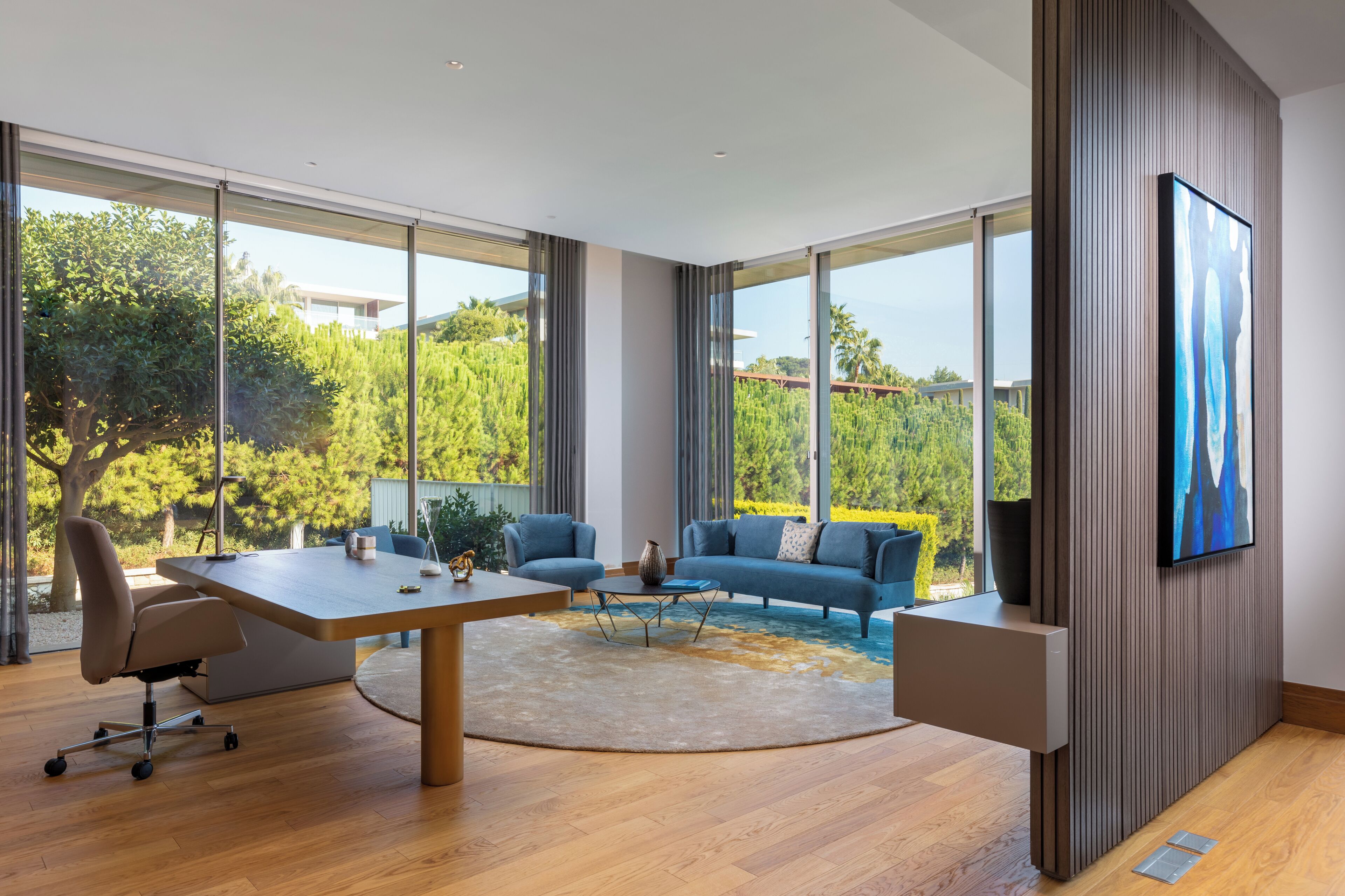 Una habitación moderna con un escritorio grande, un sofá azul y ventanas amplias que dan a vistas de zonas verdes y edificios.