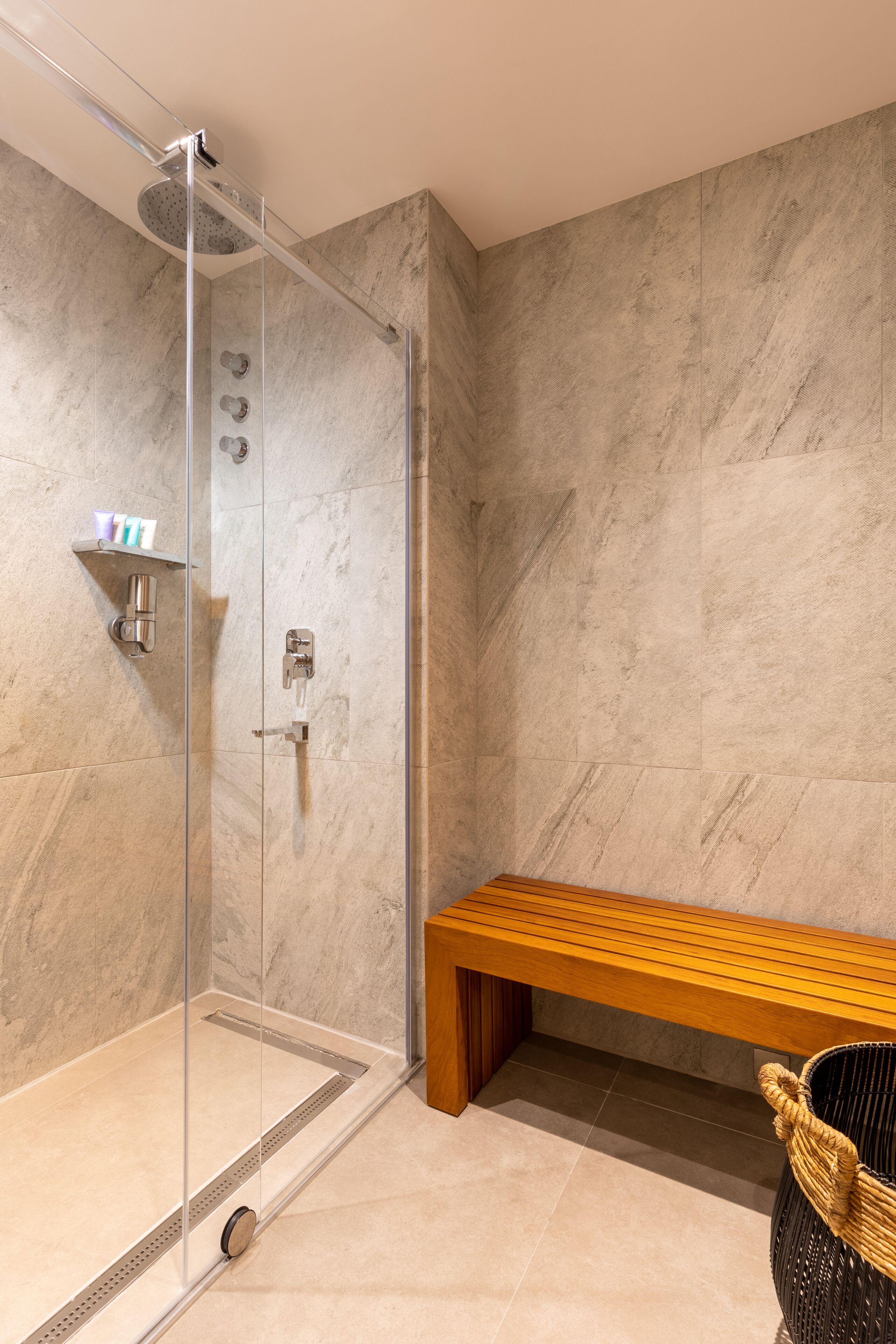 Un baño moderno con cabina de ducha de vidrio, un banco de madera y una cesta de mimbre.