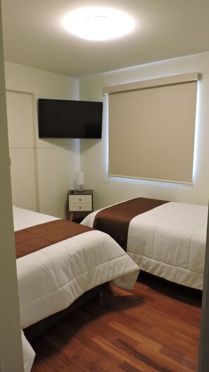 A hotel room with two beds, a flat-screen TV mounted on the wall, a bedside table with a lamp, and a window with a blind.