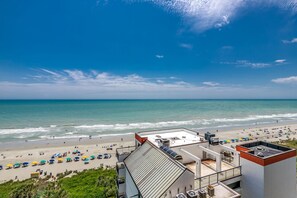 A beach view from a hotel rooftop with a clear sky, beach umbrellas, and a sandy shoreline.
