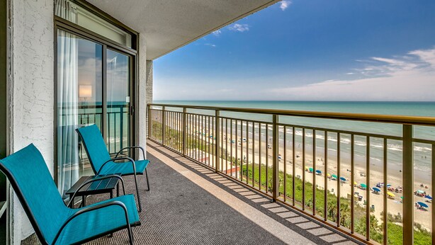 Ein Balkon mit zwei Stühlen und einem Tisch, der Blick auf einen Strand mit Sonnenschirmen und Menschen bietet.
