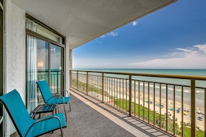A balcony with two chairs and a table, overlooking a beach with umbrellas and people.