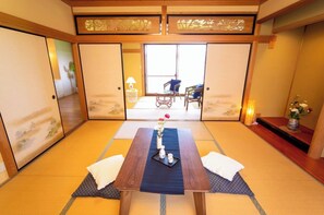 A traditional Japanese room with tatami flooring, a low wooden table with a tray, two cushions, and a view through a sliding door to a balcony with a chair and a lamp.