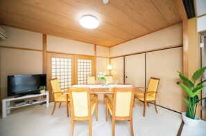 A traditional Japanese-style room with tatami flooring, a low wooden ceiling, a television on a white stand, a dining table with chairs, and a potted plant.