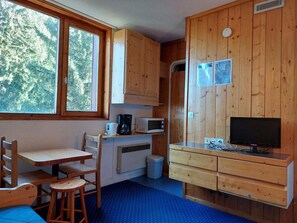 A compact kitchen with wooden cabinets, a microwave, a coffee maker, a TV, and a small dining area with chairs and a table.