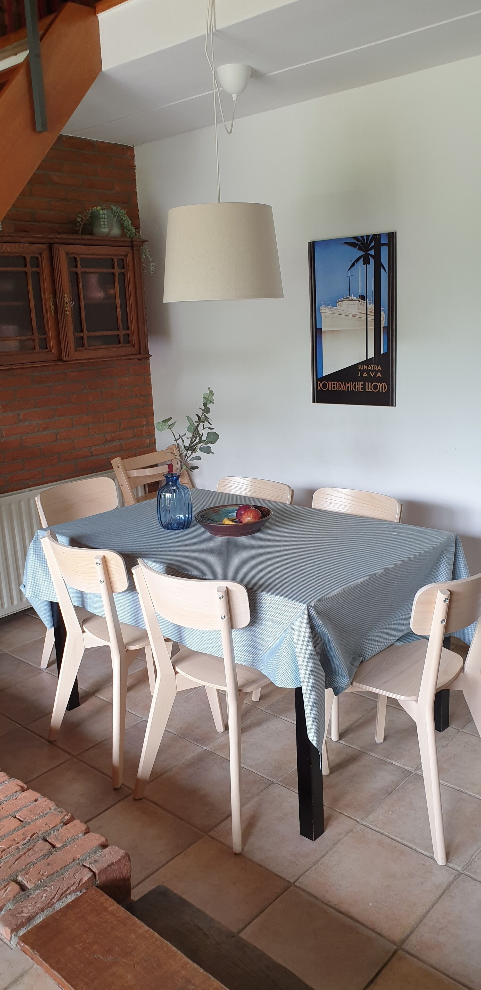 Comedor con una mesa lista para cuatro personas, rodeada de sillas de madera, un mantel azul y un florero con plantas.