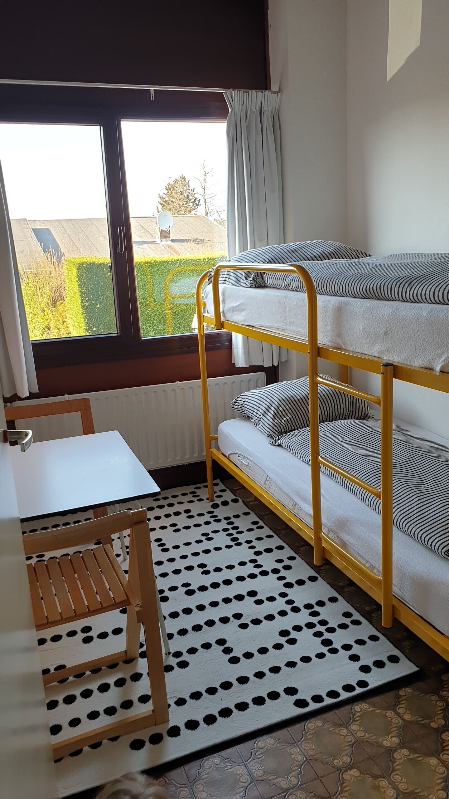 Een stapelbed met uitzicht op een tuin door een raam, een wit bureau en een vloerkleed met stippen.