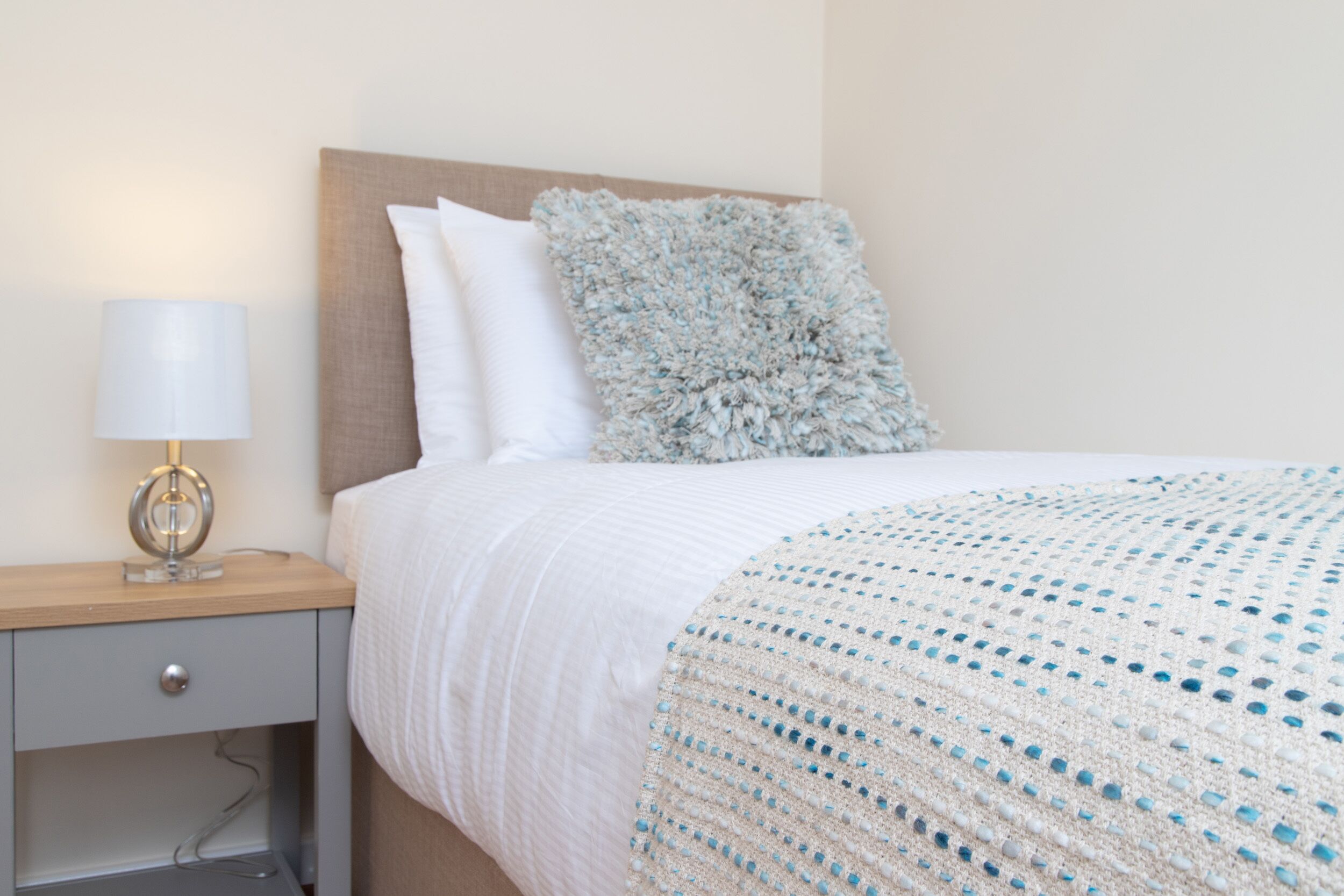 Un lit bien fait, recouvert d’une couette blanche et d’un plaid à motifs bleus et blancs. On y trouve une table de chevet en bois, avec une lampe et un tiroir.