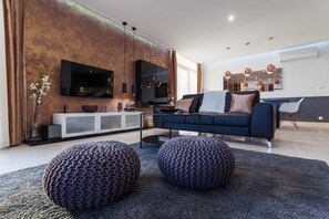 Ein modernes Wohnzimmer mit einem dunkelblauen Sofa, zwei gestrickten Poufs, einem Glastisch und einem Flachbildfernseher, der an einer strukturierten Wand befestigt ist.