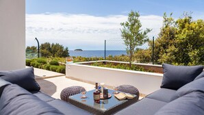 A balcony with a view of the sea, a wicker table with a bottle and glasses, and grey upholstered seating. - Luxury  Villa   Adrian (Vela Luka)