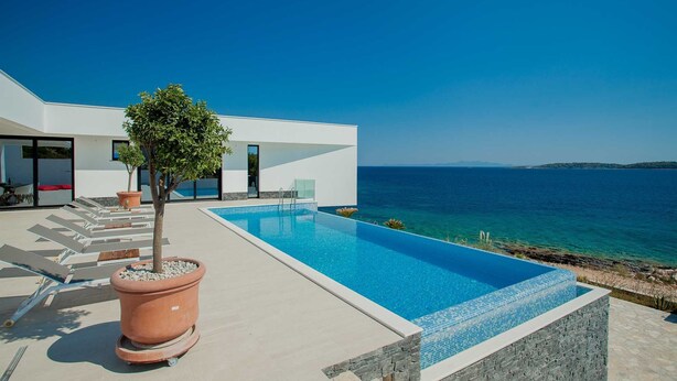 Ein moderner Poolbereich mit freiem Blick auf das Meer, einem Blumentopf mit einem Baum und einem zeitgemäßen Gebäude mit großen Fenstern.