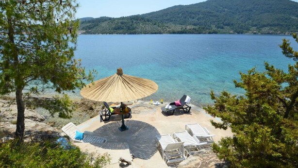 Un cadre idyllique au bord de la plage, avec un parasol en chaume, des chaises longues et une vue sur un lac paisible entouré de collines.