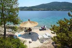 A beachside setting with a thatched umbrella, lounge chairs, and a view of a calm lake surrounded by hills.