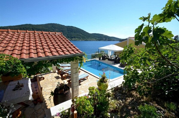 A pool area with a view of the sea and hills, a covered seating area, and a pergola with plants.