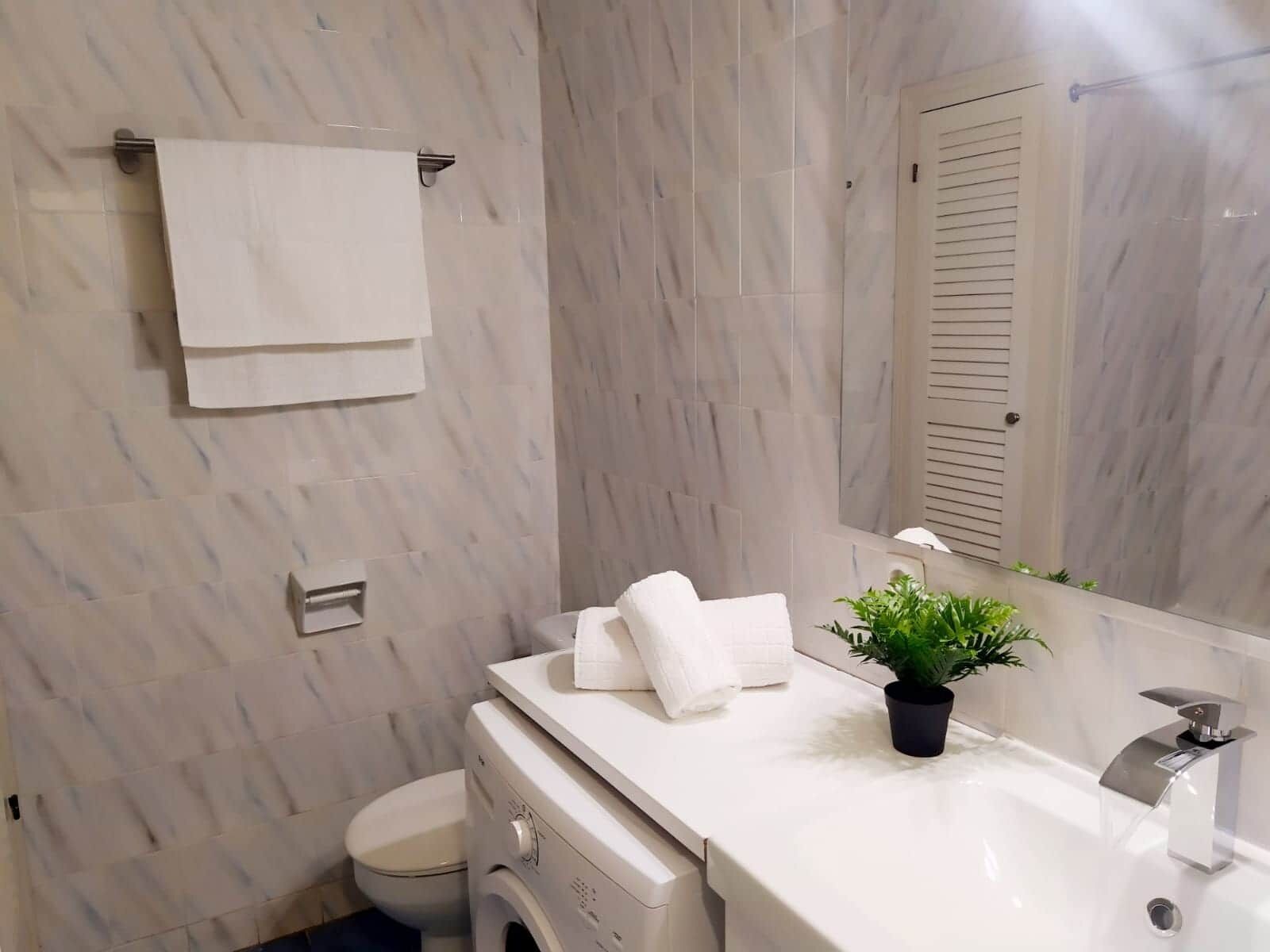 Un bagno con parete in marmo, lavandino bianco, specchio, porta asciugamani e una pianta in vaso.