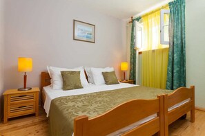 Uma cama bem arrumada com colcha estampada, duas mesas de cabeceira com abajures, um quadro na parede e uma janela com cortinas.