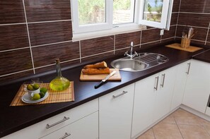 A kitchen with dark brown tiles, a window, a sink, and a cutting board with bread and a knife. - House House Čafo- Padovan (Vela Luka)