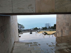 A view from a stone staircase leading to a terrace overlooking the sea, with a cityscape in the background.
