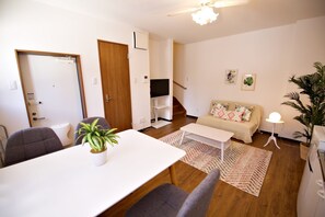 A modern living room with a white sofa, a dining table with chairs, a ceiling fan, and a TV mounted on the wall.