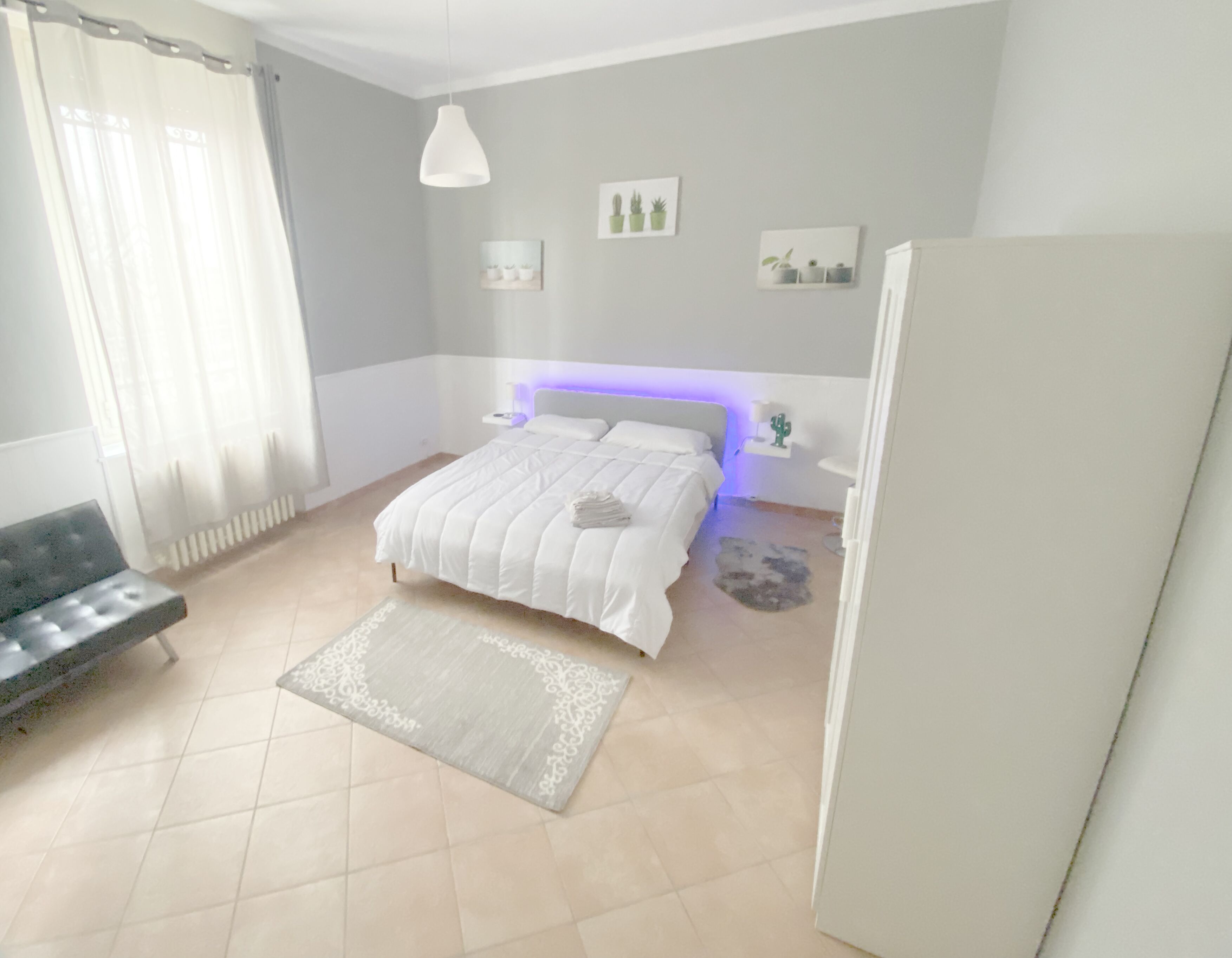 Quarto com cama, mesa de cabeceira, cadeira, radiador, janela com cortinas e parede com quadros.