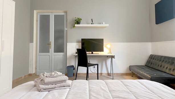 Ein Schlafzimmer mit einem Bett, einem Schreibtisch mit Computer, einem Sessel, einer Couch und einer Tür.