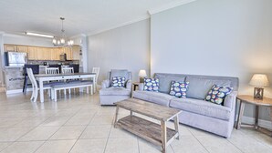 Ein modernes Wohnzimmer mit einem grauen Sofa, passenden Kissen, einem Holztisch und einem Essbereich mit weißem Tisch und Stühlen.