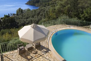 A pool area with a stone deck, a covered seating area, and a view of the sea and forested hills.