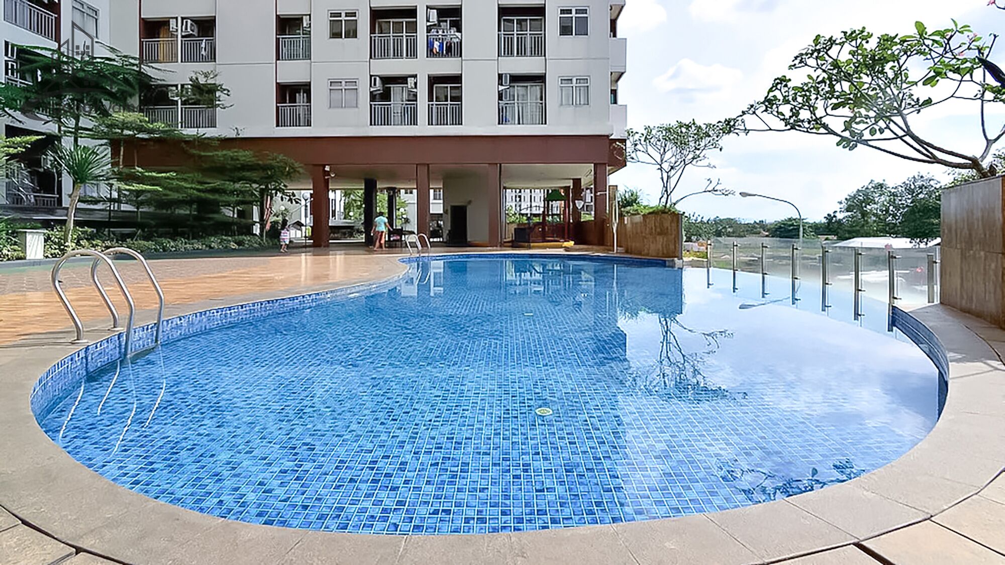 A swimming pool with a tiled edge, surrounded by a covered area with chairs and a building in the background.