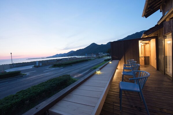 A wooden deck with chairs overlooking a road and a body of water, with mountains in the background.