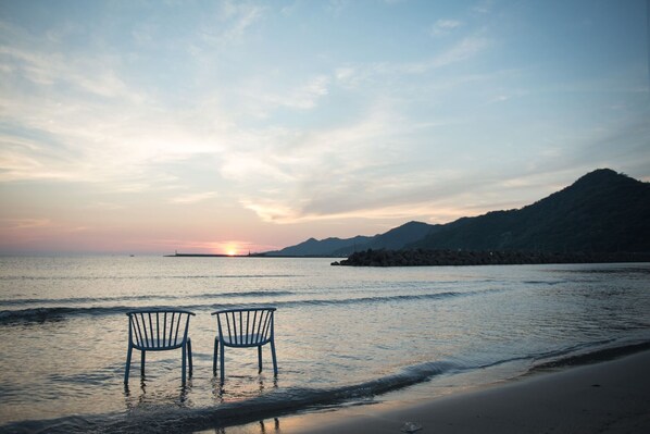 Two chairs by a calm sea at sunset with a mountain in the background.