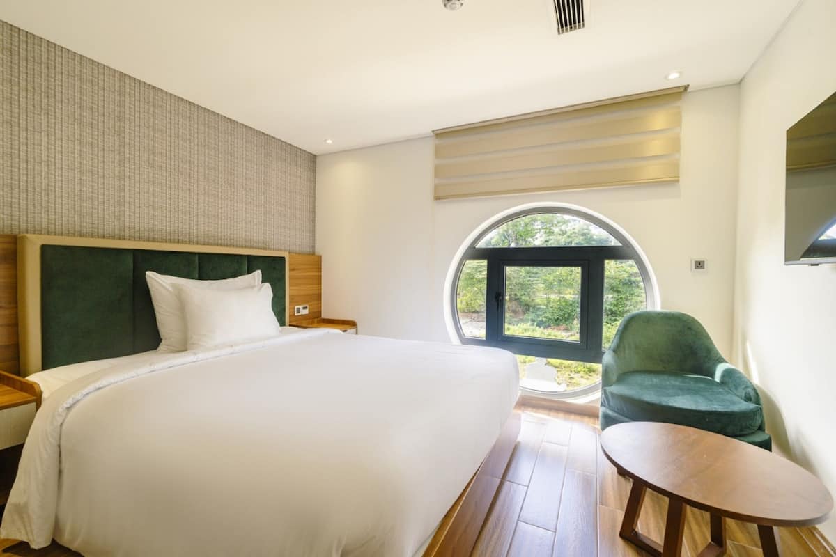 Une chambre à coucher avec un grand lit, une fenêtre ronde, un fauteuil vert et une petite table.