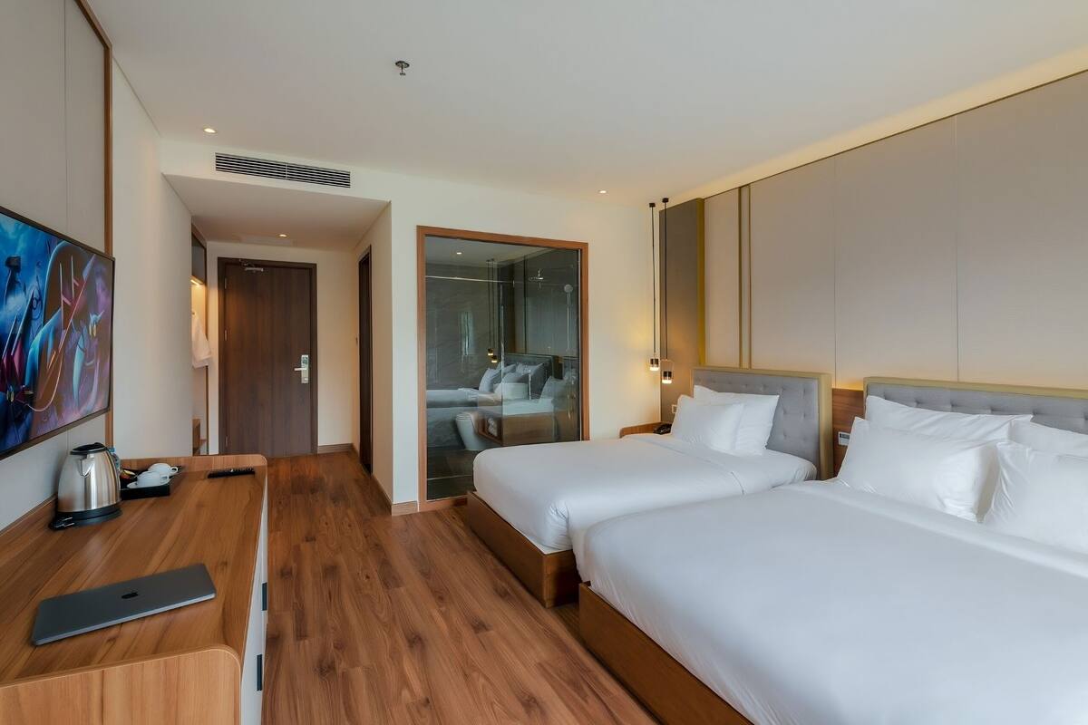 Une chambre d’hôtel avec deux lits, un bureau en bois avec un ordinateur portable, un tableau et une vue sur un balcon avec un canapé.