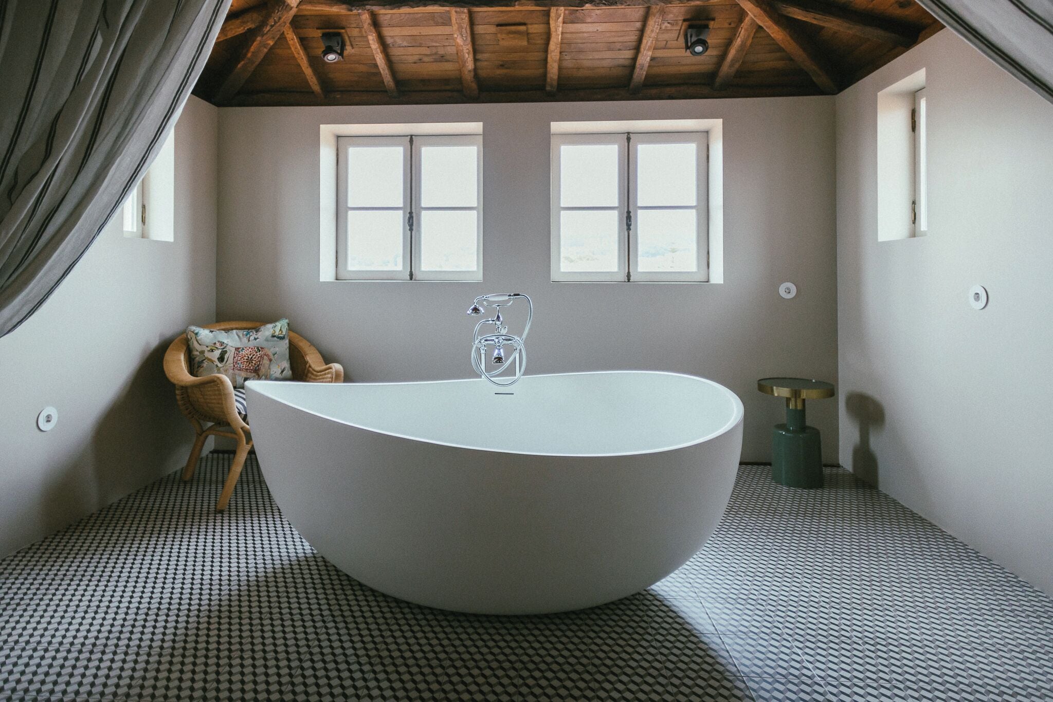Un baño moderno con una bañera independiente, dos ventanas, techo de madera y suelo estampado.