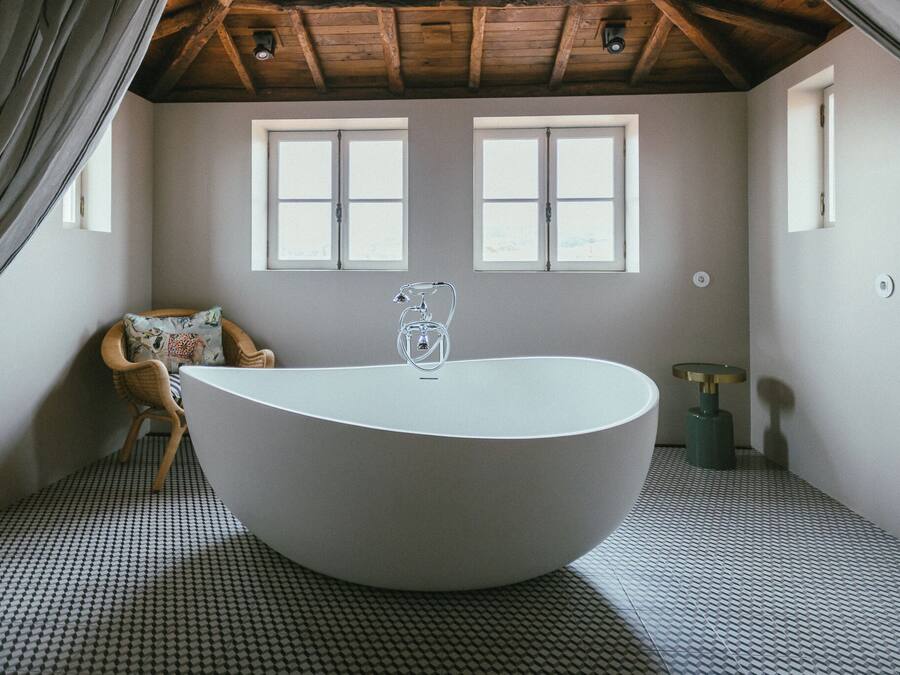 Un baño moderno con una bañera independiente, dos ventanas, techo de madera y suelo estampado.