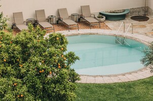 Una piscina con sillones de descanso y una pequeña mesa, rodeada por un patio de ladrillo y vegetación.
