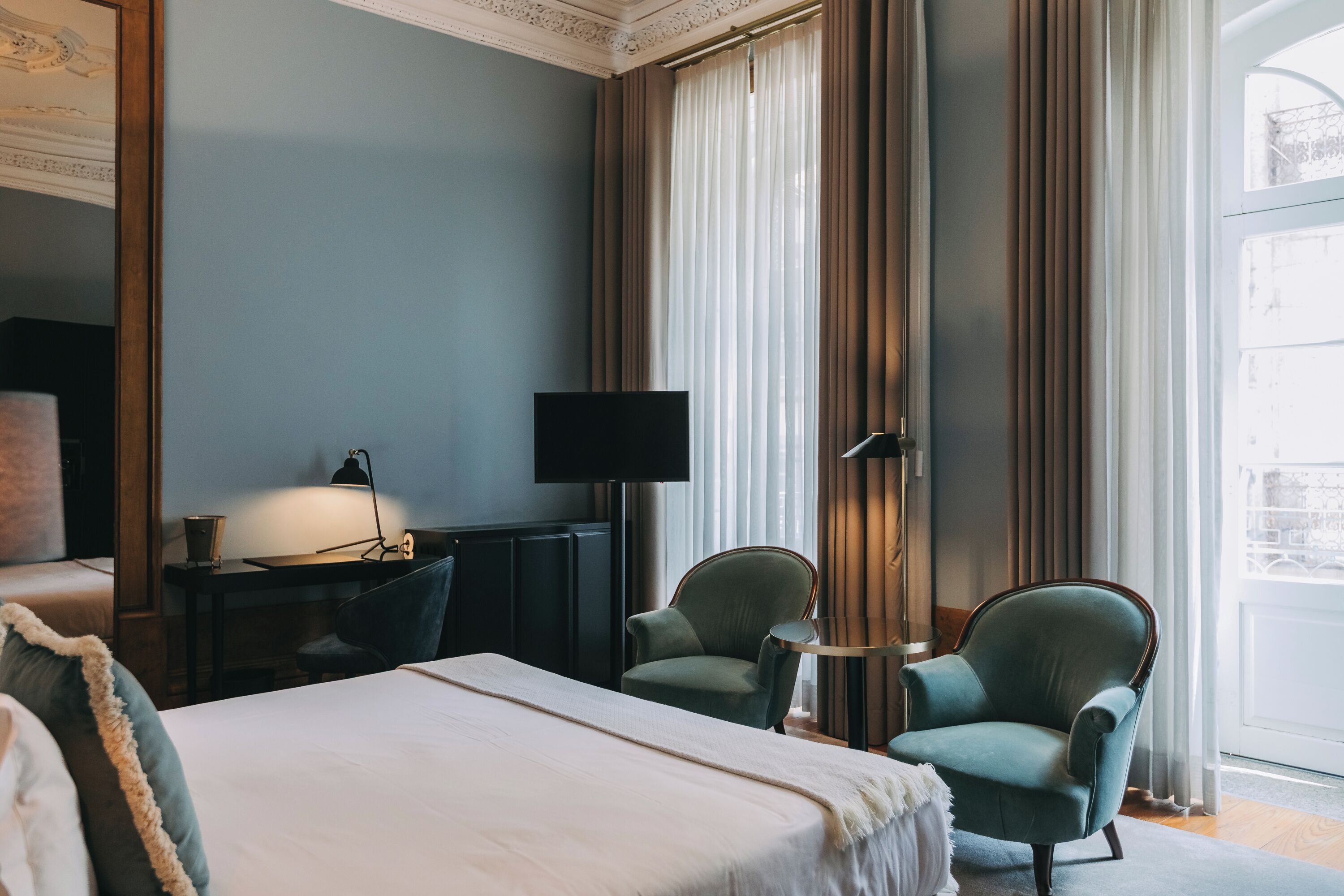 Une chambre d’hôtel avec un grand lit, deux fauteuils, un bureau avec un téléviseur à écran plat et une grande fenêtre avec des rideaux.