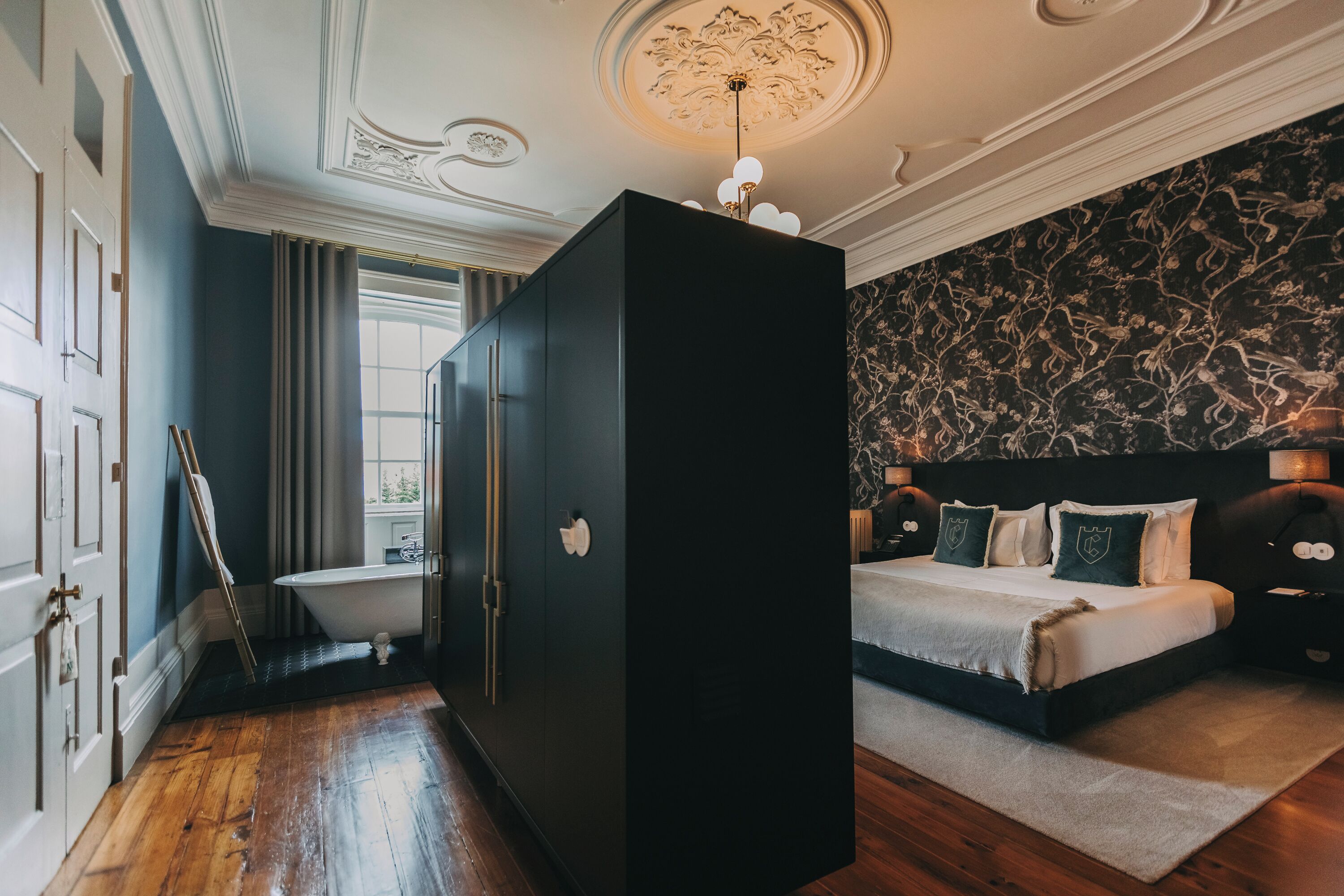 A bedroom with a large bed, a black wardrobe, a bathtub, and a window with curtains.